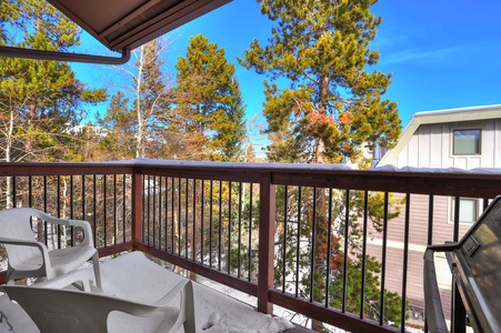 A snowy balcony with two white chairs, a wooden railing, and a clear view of nearby trees and buildings under a bright blue sky.