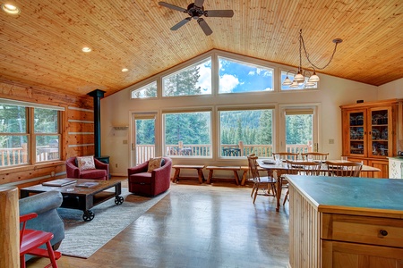 A spacious cabin interior featuring a living area with red armchairs, a coffee table, a dining area with a wooden table and chairs, large windows offering forest views, and a light wooden ceiling.