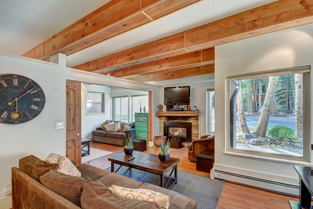 A cozy living room with wooden beams, a large wall clock, a fireplace, and a TV. It features brown sofas, a wooden coffee table, and large windows that let in natural light.