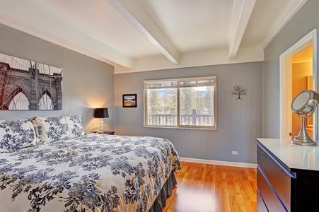 A bedroom featuring a large bed with floral bedding, a bedside table with a lamp, a dresser with a mirror, and a window with blinds. Decor includes a framed bridge photo and a tree wall decal.