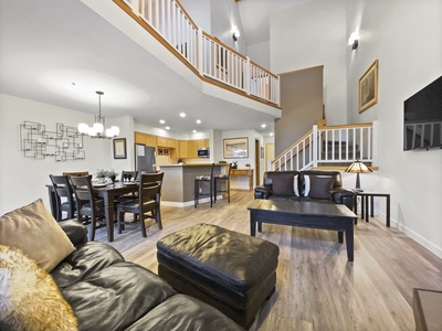 Spacious living area with modern furniture, adjacent dining table, and open kitchen. Loft and staircase are visible in the background, with high ceilings and bright lighting.