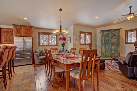 A dining room with a rectangular wooden table set for six, an ornate green cabinet, stainless steel refrigerator, and wooden bar with stools. The room has hardwood floors and multiple windows.