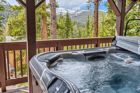 A hot tub on a wooden deck with a view of a forest and snow-capped mountains in the background.