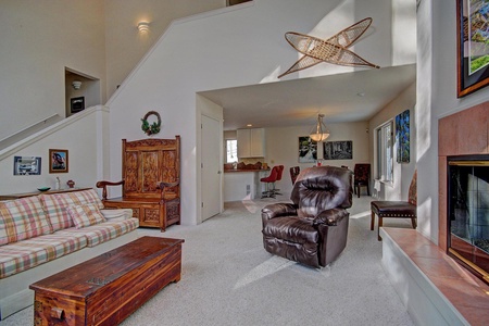 A living room with a brown recliner, plaid sofa, wooden chest coffee table, ornate cabinet, and high ceilings. The room is well-lit with natural light and features a staircase and open kitchen area.