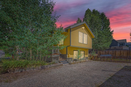 A yellow two-story house with a gravel driveway, surrounded by trees, under a colorful sunset sky.