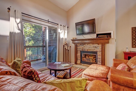 Cozy living room with leather sofas, a fireplace, wall-mounted TV, and sliding glass doors leading to a balcony with outdoor furniture. The room is decorated with colorful cushions and a striped rug.