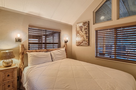 A cozy bedroom with a double bed, wooden blinds on the windows, a bedside lamp on a small wooden nightstand, and a framed artwork on the wall. Light fixtures are mounted above the headboard.