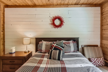 A cozy bedroom with a wooden wall, a bed adorned with plaid and striped cushions, a red wreath above the headboard, a bedside table with a lamp, and a box of tissues.