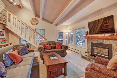 A cozy living room with a fireplace, flat-screen TV, several couches, and wooden coffee table. Large windows and a staircase leading to the upper level are visible.