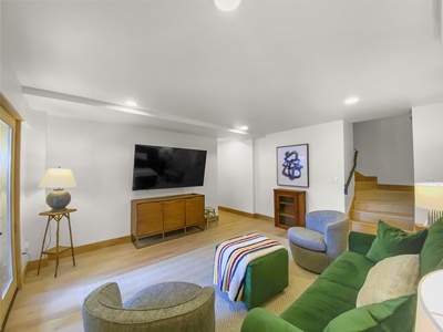 Modern living room with green sofa, armchairs, and a striped throw blanket. A large flat-screen TV is mounted on the wall above a wooden console. Room features a carpeted floor and wood accents.