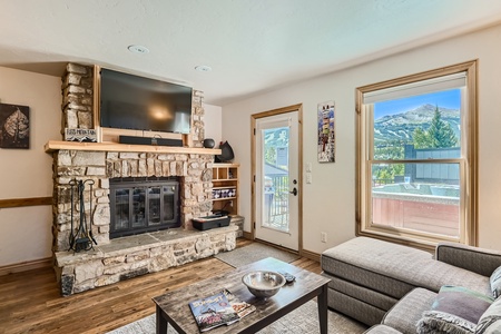 A cozy living room featuring a stone fireplace with a mounted TV, a grey sectional sofa, wooden coffee table, and bright windows showcasing a scenic mountain view.