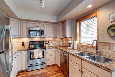 A modern kitchen with stainless steel appliances, granite countertops, and light-colored cabinets. There is a double sink under a window with wooden blinds, and the floor is hardwood.