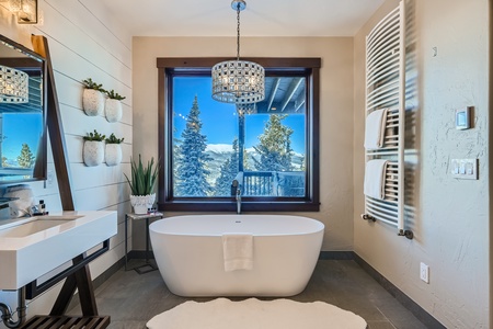 A modern bathroom with a freestanding tub under a chandelier, large window with snowy mountain view, and wall-mounted towel racks.