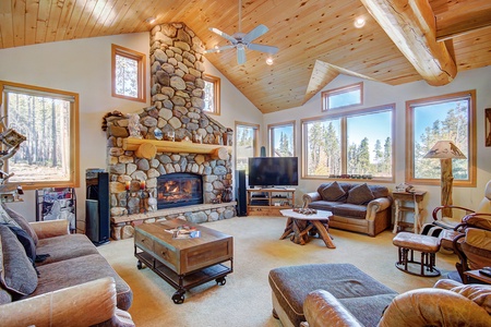 A cozy living room with a wooden ceiling, stone fireplace, and large windows. It contains sofas, a TV, a coffee table, and various chairs. View shows a forested landscape outside.