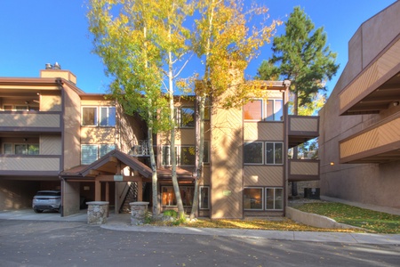 A three-story apartment building with balconies and a covered entrance. There are trees in front and a parked car in a partially visible carport to the left.