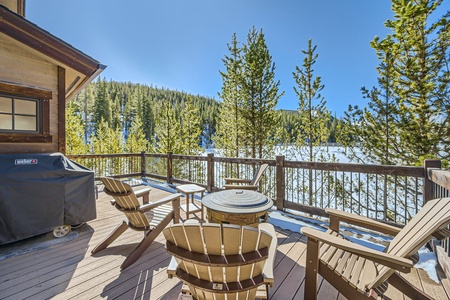 Deck with Adirondack chairs around a fire pit, a covered grill on the left, overlooking a snowy forest and hills under a clear blue sky.