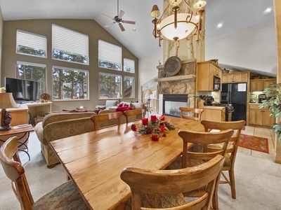 Open-concept living and dining area with high ceilings, large windows, a stone fireplace, wooden furniture, and a modern kitchen in the background.