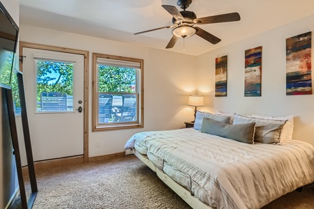 A bedroom with a bed, nightstand, lamp, ceiling fan, artwork on the wall, and a door and window leading outside, with trees visible through the glass.