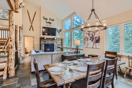 A rustic lodge living area with a dining table set for six, a gray sectional sofa, a stone fireplace, large windows, wooden beams, and "Twin Creek Lodge Est. 2017" on the wall above the fireplace.