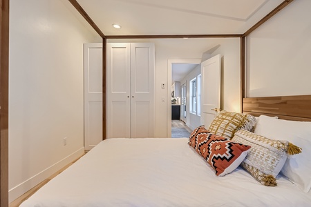 A small bedroom with a white bed, multiple patterned pillows, a wooden headboard, white closets, and a door leading to an adjacent room.