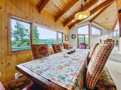 A dining room with a long wooden table covered in a patterned tablecloth, surrounded by upholstered chairs, and large windows providing a view of trees and mountains.