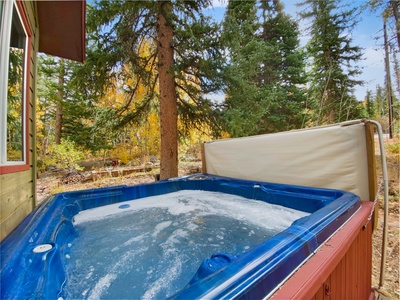 A blue hot tub filled with bubbling water is situated outdoors next to a wooden cabin, surrounded by tall trees and forest foliage.