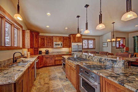 A spacious kitchen with wooden cabinets, granite countertops, stainless steel appliances, and a central island. Hanging pendant lights and ample natural light from large windows enhance the space.