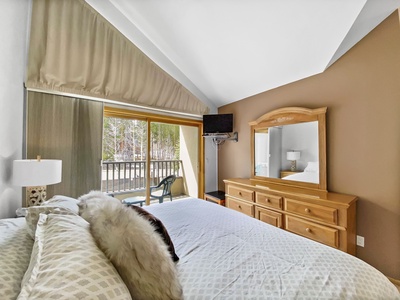 A bedroom with a bed, dresser, mirror, TV, and sliding glass door leading to a balcony with outdoor chairs. The room has vaulted ceilings and a large window with a forest view.