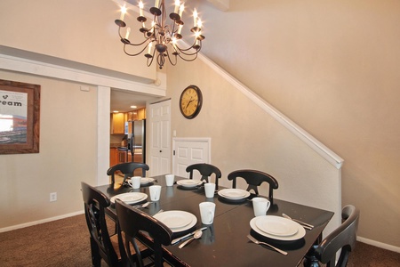 A dining room with a black table set for six, featuring white dishes and cups. A black chandelier hangs above the table, with a view of a kitchen and a staircase adjacent.