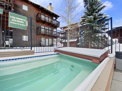 A hot tub with a partially open cover is situated outdoors, surrounded by a black metal fence. Snow is visible on the ground and on roofs of nearby wooden buildings. A sign reads "Tyra Summit II Spa House.