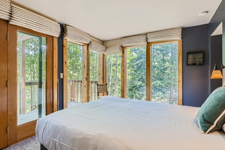 A bedroom with a large bed, blue accent wall, and windows on three sides offering a view of surrounding trees. The room features a ceiling light and a chair near the windows.