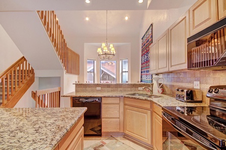 A kitchen with granite countertops, wooden cabinets, an electric stove, a dishwasher, a microwave, a chandelier, and a staircase with wooden railings in the background.