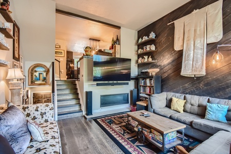 A cozy living room with a TV mounted above a fireplace, a corner couch, a coffee table, and eclectic decor, including a wall-mounted scroll and shelves filled with various items.