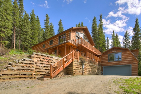 A large wooden mountain cabin with a two-car garage, elevated porch, and stairs, set against a backdrop of tall evergreen trees and blue skies.