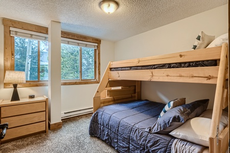 A bedroom with a wooden bunk bed, featuring a lower double bed and an upper single bed. There is a nightstand with a lamp beside the window, and the walls are painted white.