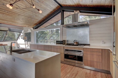 Modern kitchen with wooden cabinetry, stainless steel appliances, a large island with a sink, and a slanted wooden ceiling. Large windows offer ample natural light and outdoor views.