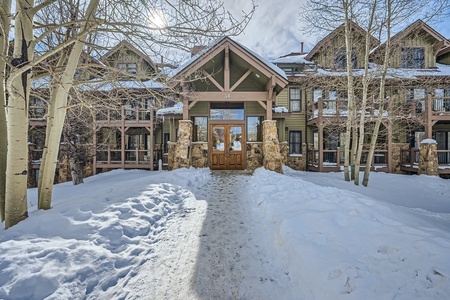 Front view of a multi-story wooden building in a snowy landscape. A path is cleared of snow leading to a large wooden door with stone pillars, surrounded by leafless trees.