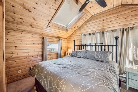 A cozy bedroom with wood-paneled walls and ceiling, a large bed with gray bedding, a skylight, and a small window with curtains.