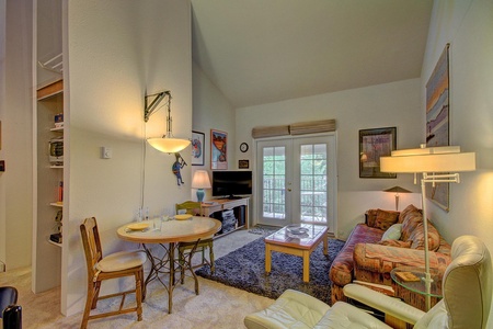 A cozy living room and dining area with a round table, chairs, a sofa, a coffee table, TV, and large glass doors leading to an outdoor area. The room is well-lit with natural and artificial light.