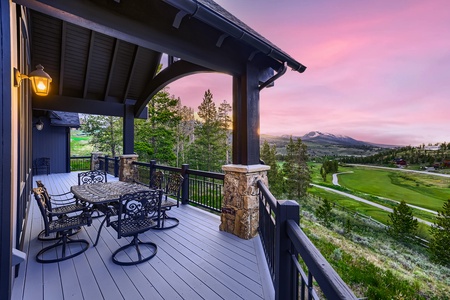 A covered outdoor deck with a dining table and chairs overlooks a lush golf course and mountain landscape under a pink sunset sky.