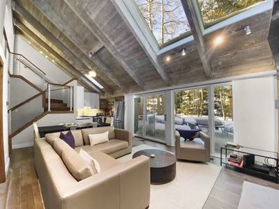 A modern living room with a vaulted, wooden ceiling, large skylights, a sectional sofa, an armchair, a round coffee table, and sliding glass doors opening to a patio with outdoor furniture.