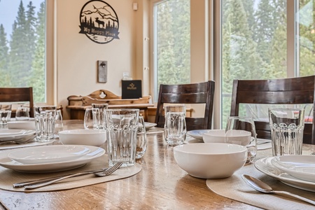 A dining table is set with white plates, bowls, glasses, and silverware. Wooden chairs surround the table. Large windows reveal a forested view outside.