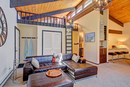 A cozy living room features a large leather sectional sofa, a wooden coffee table, a staircase leading to a loft, a modern chandelier, and a small kitchen area with bar stools in the background.