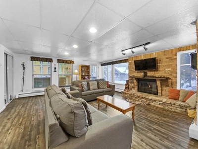 A living room with two gray sofas, a coffee table, and a brick fireplace with a TV on the mantle. The room has wooden flooring, large windows, and a built-in shelf with decor.