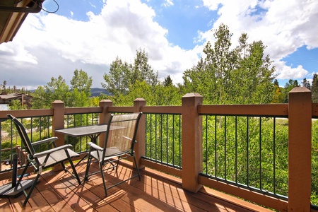 A sunny balcony with a table and four chairs overlooks a lush green landscape with tall trees and a partly cloudy sky.