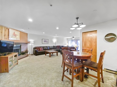 A cozy living room with a fireplace, sectional sofa, TV, round dining table with four chairs, and modern light fixtures. Wall art and a glass sliding door leading to a balcony are visible.