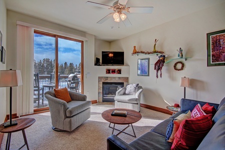 A cozy living room with a sofa, two armchairs, a coffee table, a TV mounted above a fireplace, and large windows with a view of a snowy landscape. The room is decorated with colorful pillows and wall art.