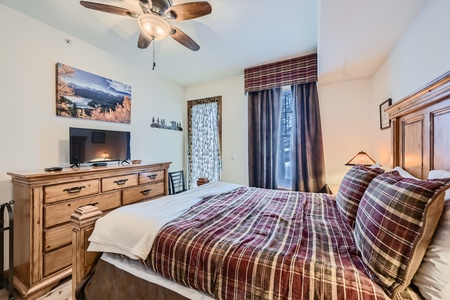 A cozy bedroom with a ceiling fan, plaid bedspread, wooden dresser with TV, nature painting, and tall window with curtains.