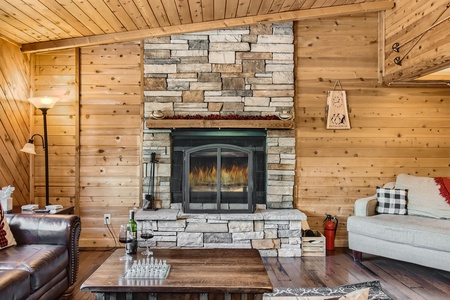 A cozy living room with a stone fireplace, wooden walls, and a wooden ceiling. A brown leather chair, gray sofa, and wooden coffee table with a wine bottle and glasses are in the foreground.