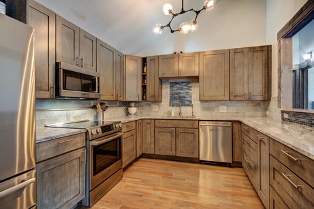 A modern kitchen with wood cabinets, stainless steel appliances, a tile backsplash, and a central light fixture. The kitchen includes a refrigerator, oven, microwave, and dishwasher.